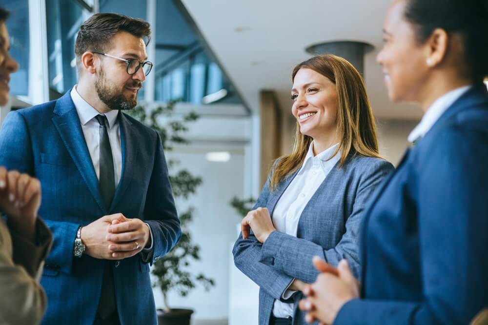Four business professionals converse in a bright modern office, with a smiling woman and a bearded man in suits prominent in the center.