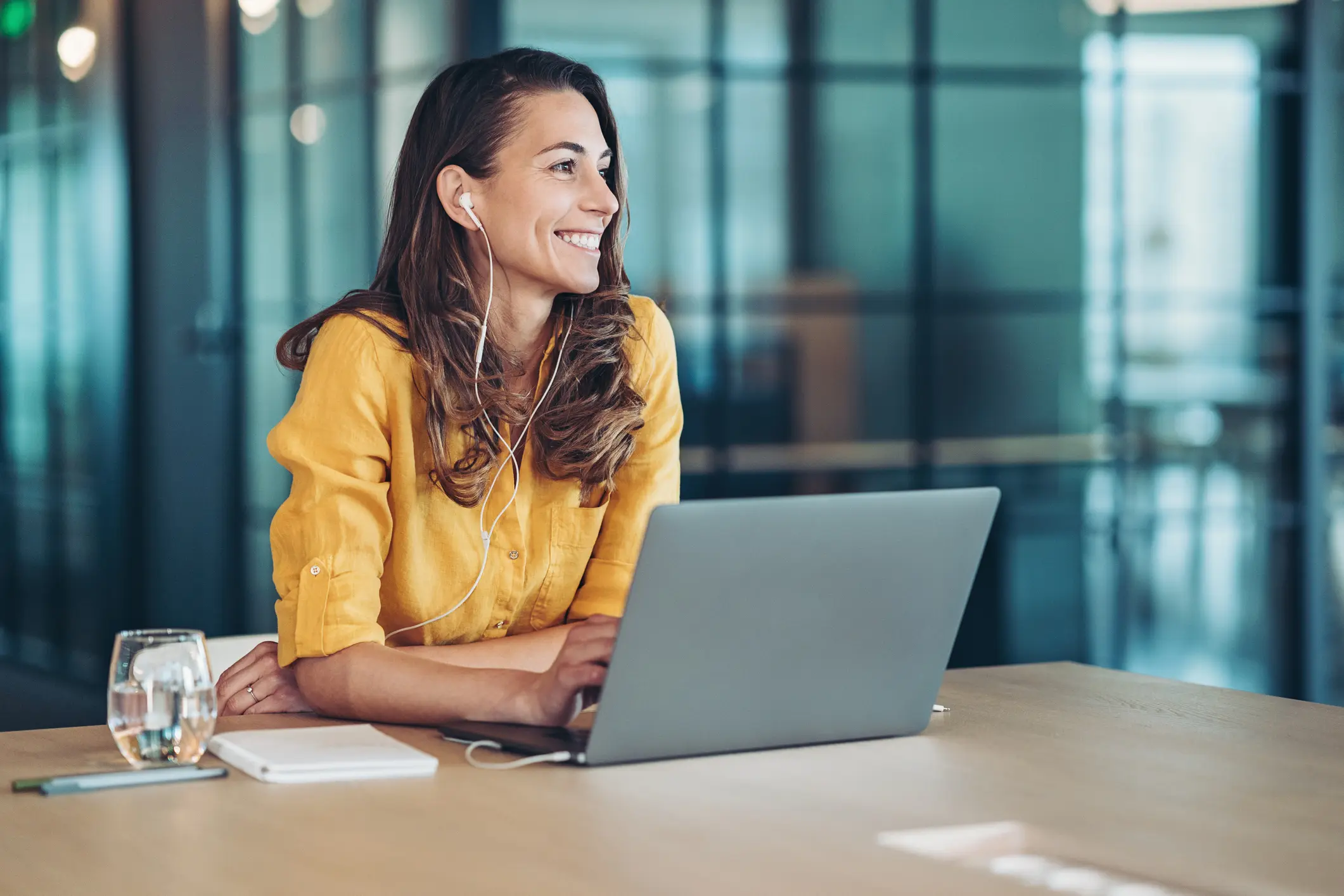 Smiling woman in a yellow shirt wearing wired earbuds and sitting behind an open laptop at a table.
