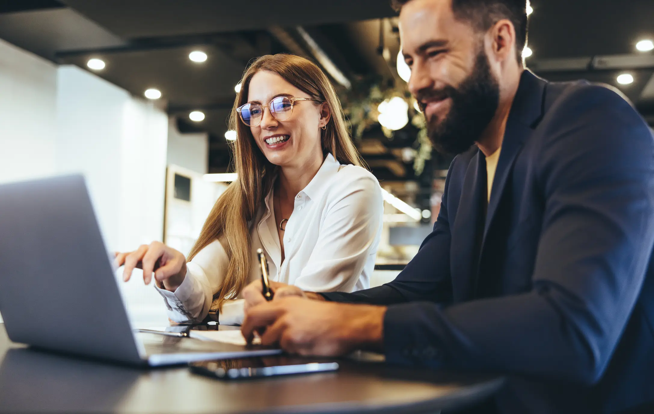 Two smiling colleagues collaborate at a table with an open laptop, papers, and a tablet.