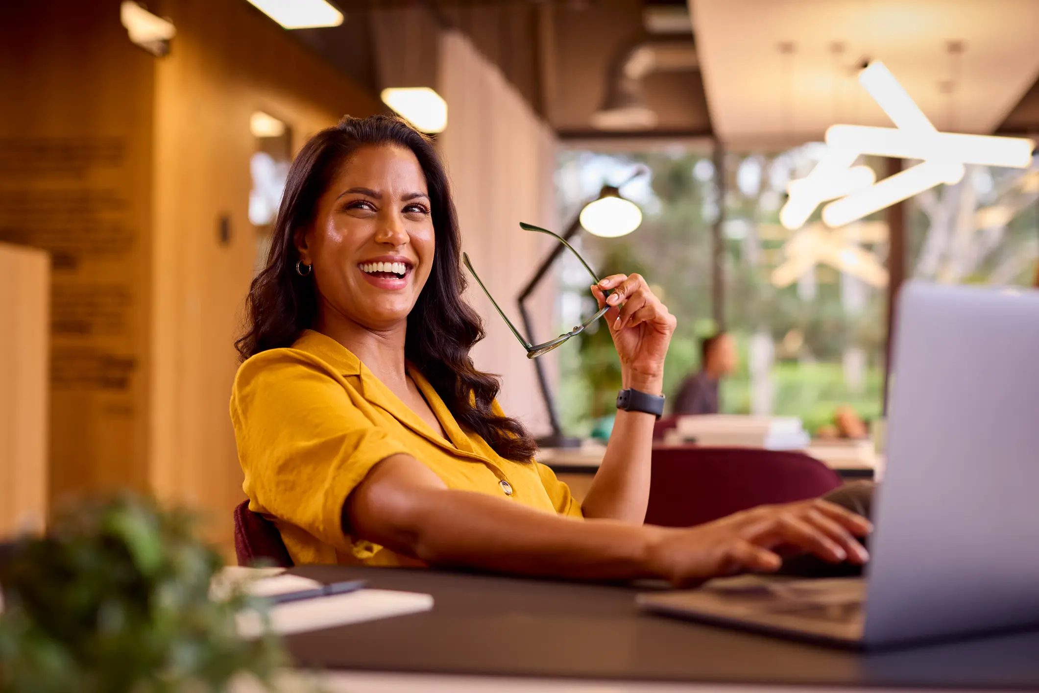 Smiling woman in a yellow blouse holds eyeglasses while working at a laptop in a warmly lit workspace.