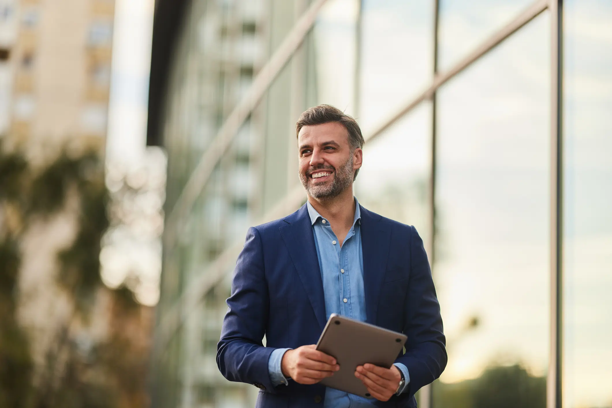 Smiling man in a navy blazer holding a tablet beside a sunlit glass wall.