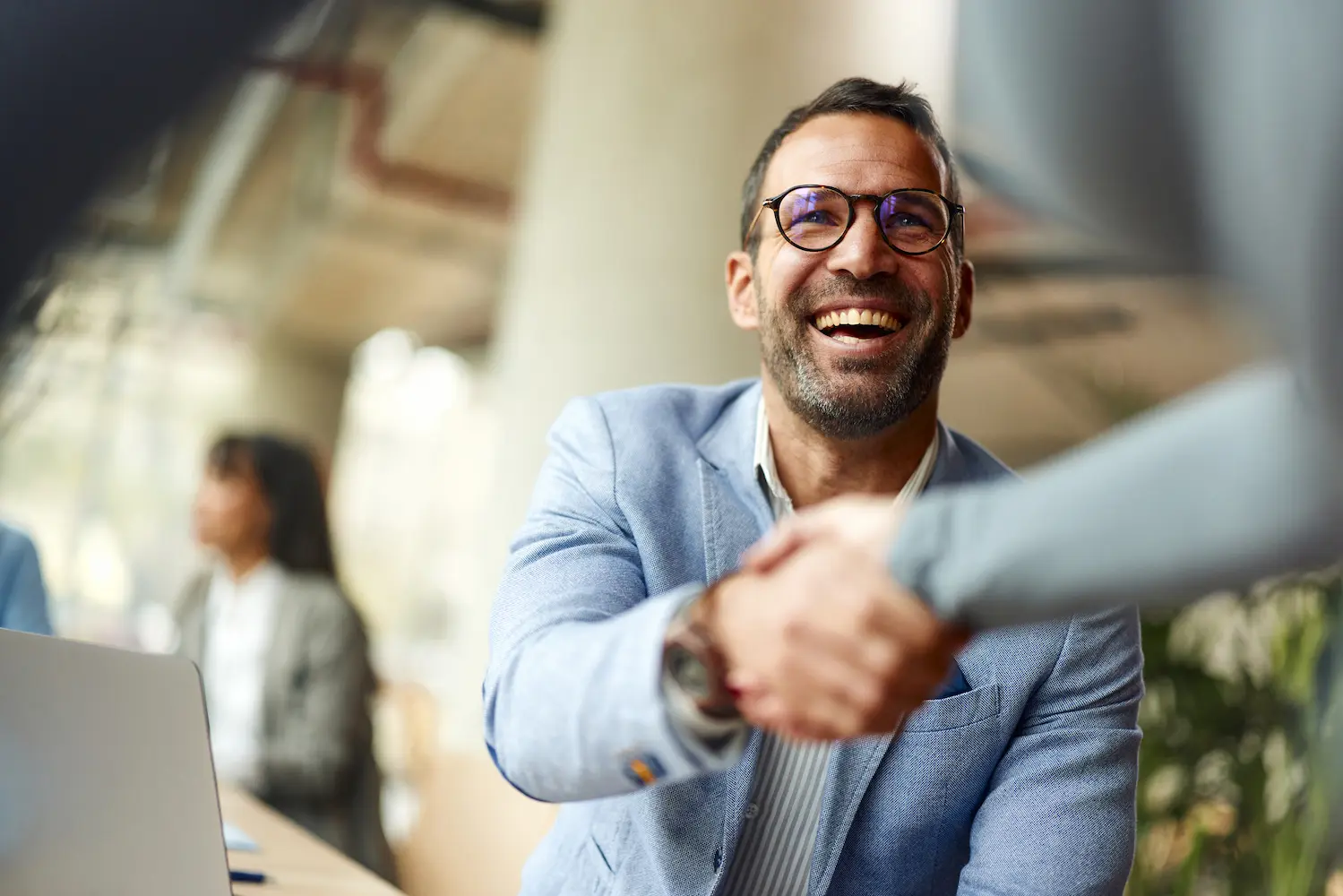 Smiling man with glasses and a salt-and-pepper beard, wearing a light blue blazer, shaking hands with a person in the foreground in a bright, modern office. A blurred colleague and laptop appear in the background.