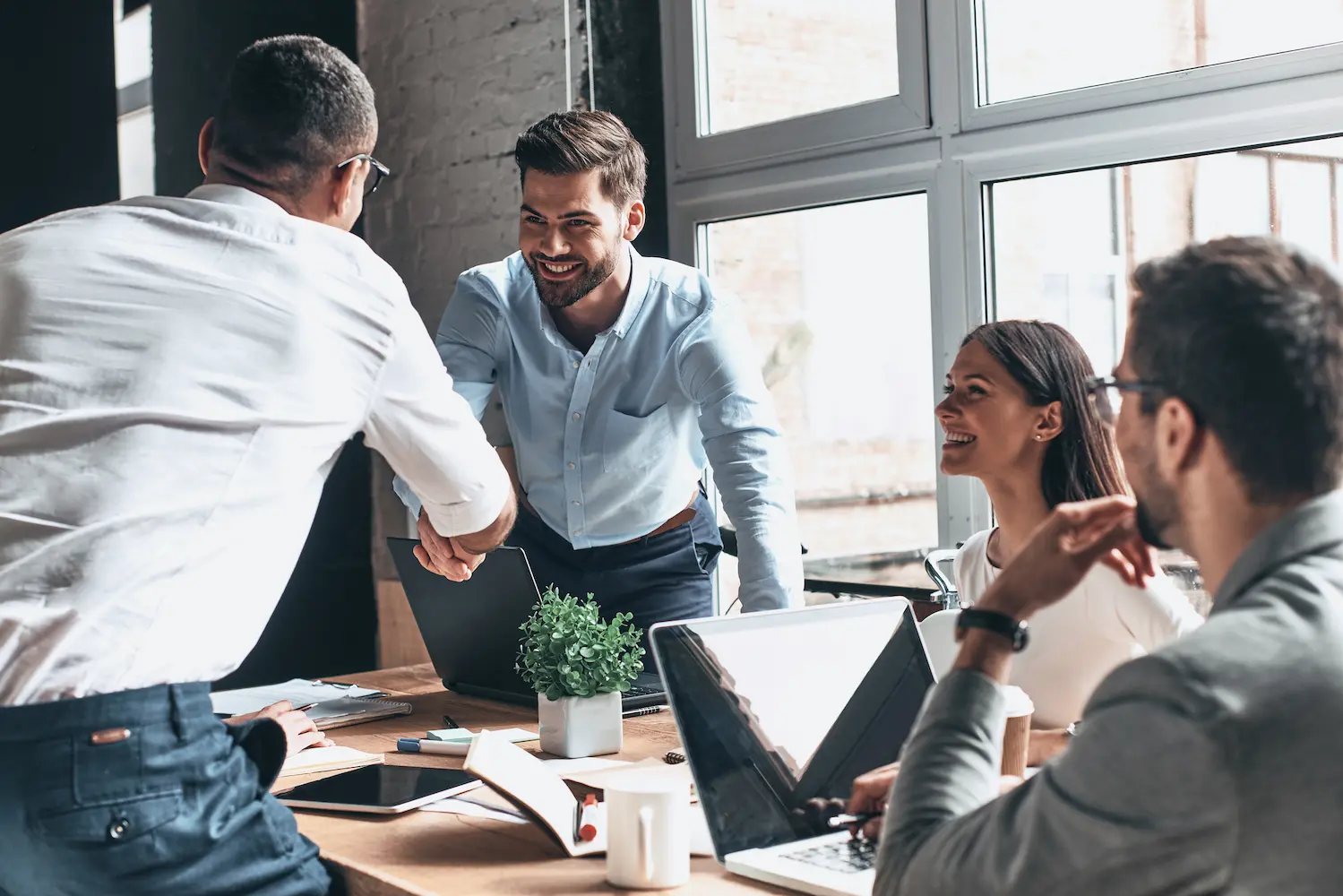 Four coworkers meet around a table as two men shake hands in a bright office.